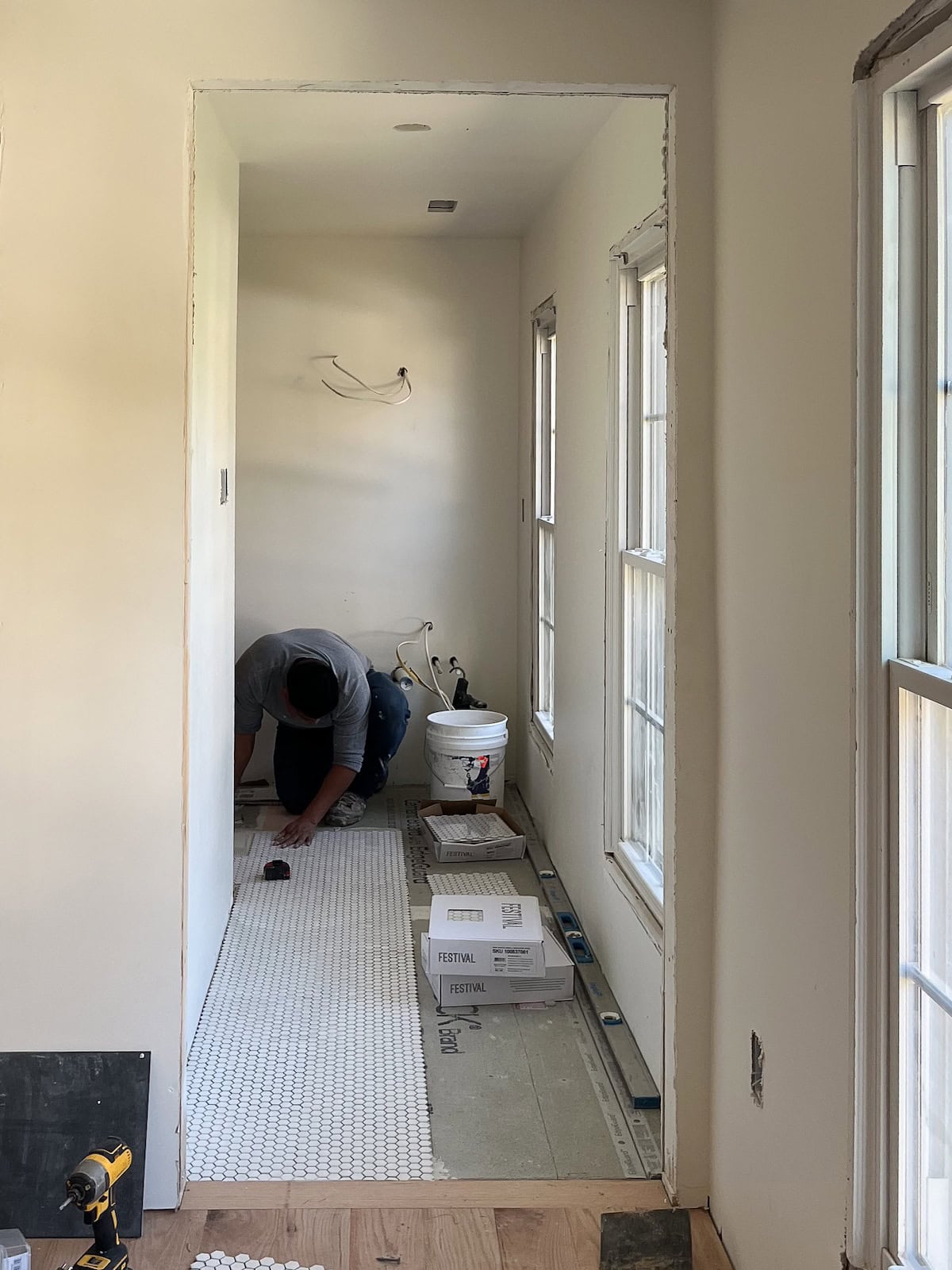 A tile installer measuring and laying out white mosaic floor tile in a residential bathroom.